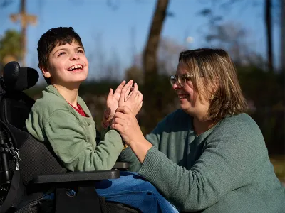 A boy in a wheelchair smiling while his caregiver playing with him outdoor