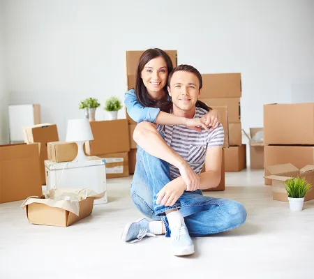 A couple sitting down on the floor looking at the camera and smiling, behind unpacked boxes